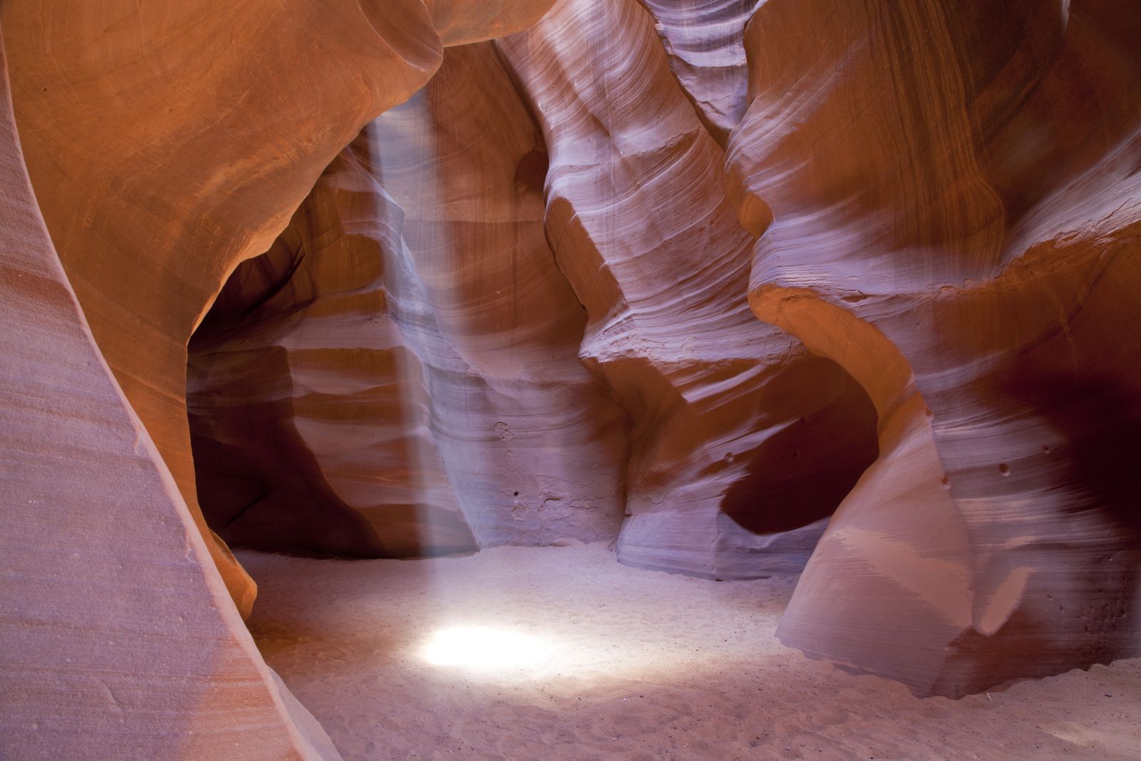A shaft of sunlight falling to the sandy floor of a narrow orange slot canyon